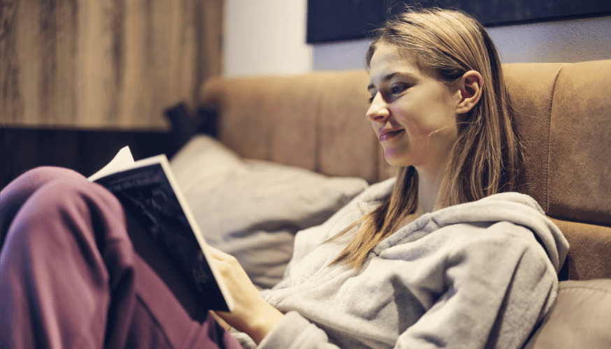 Eine Frau in einem grauen Kapuzenpullover sitzt auf einem Sofa, liest ein Buch und lächelt.