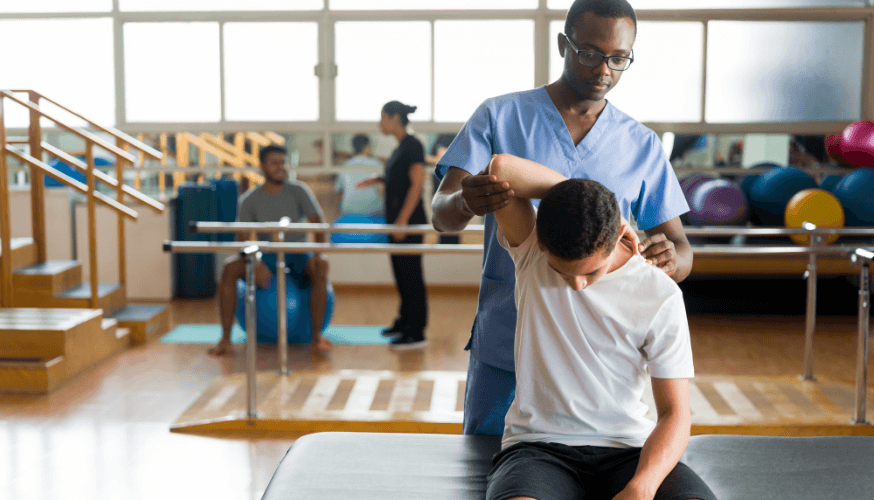 Physiotherapeut in blauer Uniform führt eine Schulterübung mit einem jungen Patienten in einem hellen Therapieraum durch, weitere Personen im Hintergrund.