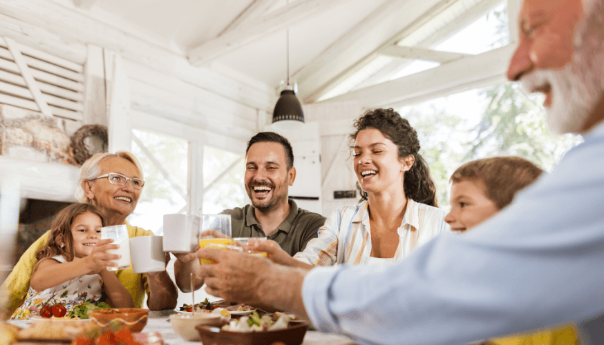 Eine Gruppe von Menschen sitzt an einem Tisch, lächelt, erhebt ihre Gläser und genießt gemeinsam eine Mahlzeit im Innenbereich.