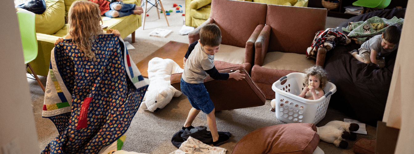 Kinder spielen in einem überfüllten Wohnzimmer mit Kissen, einer Decke und einem Wäschekorb.