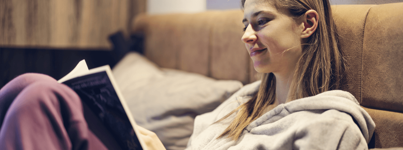 Eine junge Frau sitzt gemütlich auf einem Sofa und liest ein Buch, dabei lächelt sie leicht