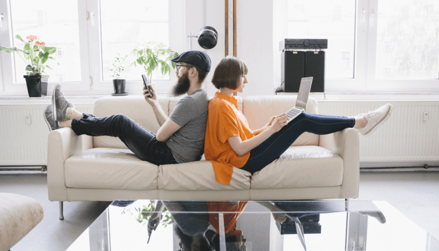 Zwei Personen sitzen Rücken an Rücken auf einem Sofa und blicken in entgegengesetzte Richtungen. Einer benutzt ein Smartphone, der andere einen Laptop. Im Hintergrund sind Pflanzen und Fenster zu sehen.