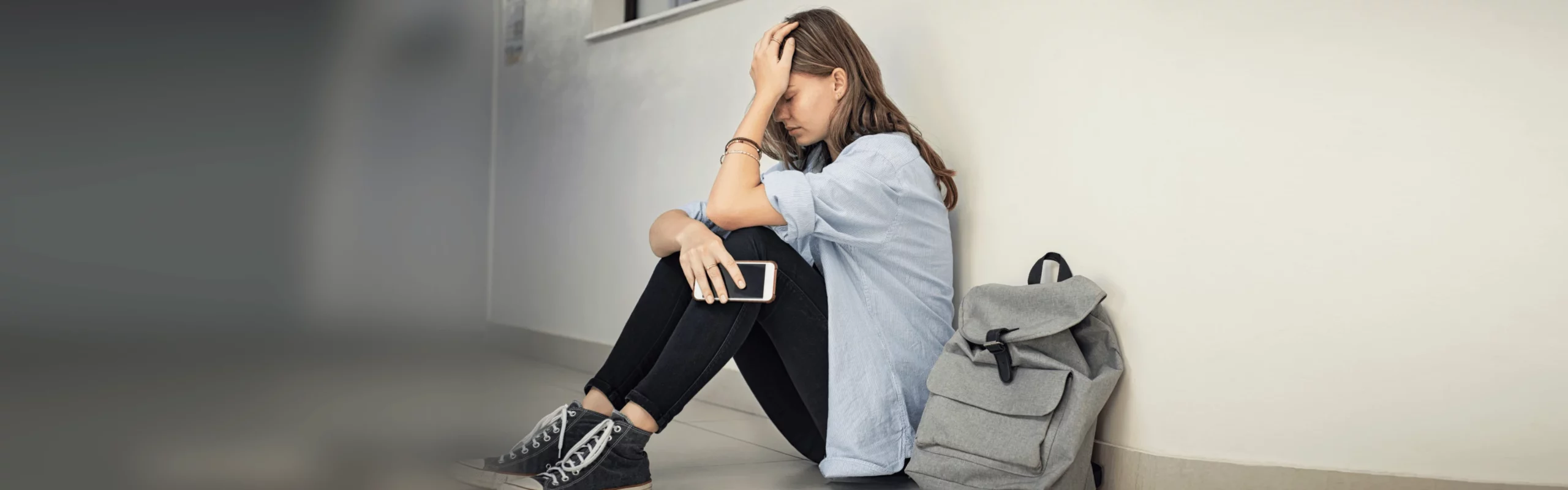 Eine Person sitzt auf dem Boden an einer Wand, hält mit einer Hand ihren Kopf und in der anderen ein Smartphone. Neben ihr steht ein grauer Rucksack.