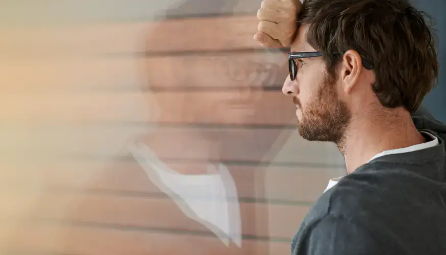 Ein Mann mit Brille lehnt seine Stirn an ein Fenster und wirkt nachdenklich oder gestresst. Sein verschwommenes Spiegelbild ist im Glas zu sehen. Der Hintergrund ist in warmen Tönen sanft unscharf.