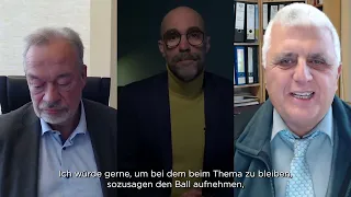 Es werden drei Männer in getrennten Videoanrufrahmen gezeigt. Der Mann auf der linken Seite blickt nach unten, der Mann in der Mitte blickt nach vorne, trägt eine Brille und einen Bart, und der Mann auf der rechten Seite lächelt. Deutsche Untertitel sind am unteren Rand zu sehen.